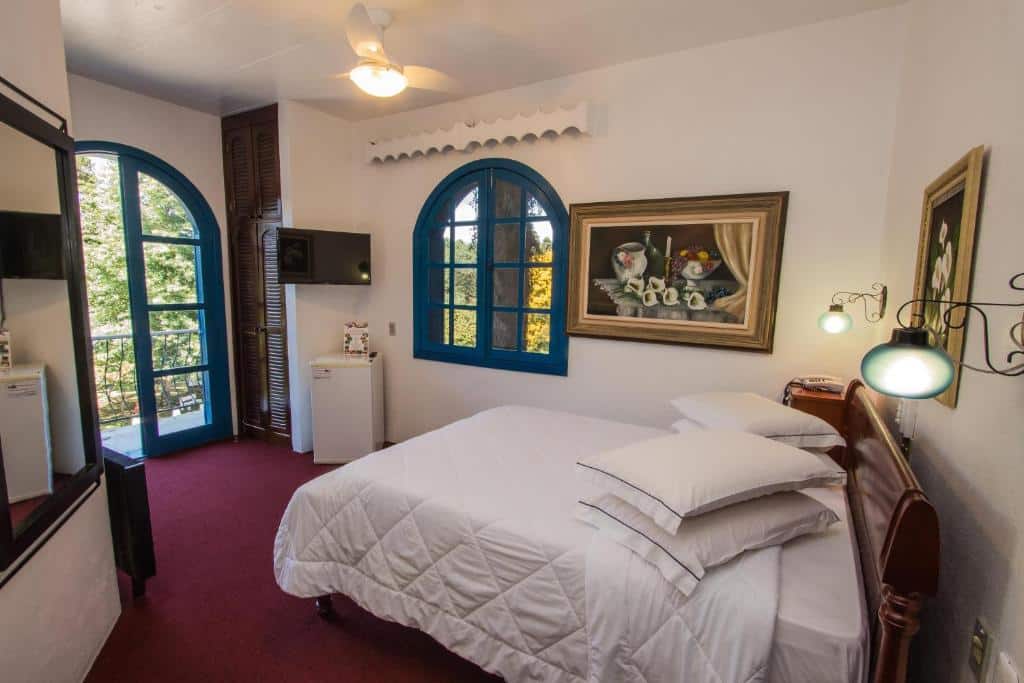 suíte do Hotel Gramado Palace com uma cama antiga de casal, janelas e portas da sacada pintadas de azul escuro, um pequeno frigobar perto da janela e dois quadros de flores brancas pendurados às paredes