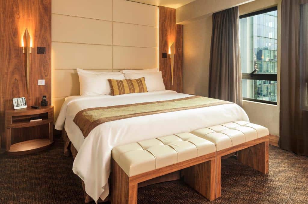 Quarto do Hotel Regal Pacific com cama de casal no centro do lado esquerdo da cama cômoda de madeira e no pé da cama bancos de madeiras estofados de tecido branco.