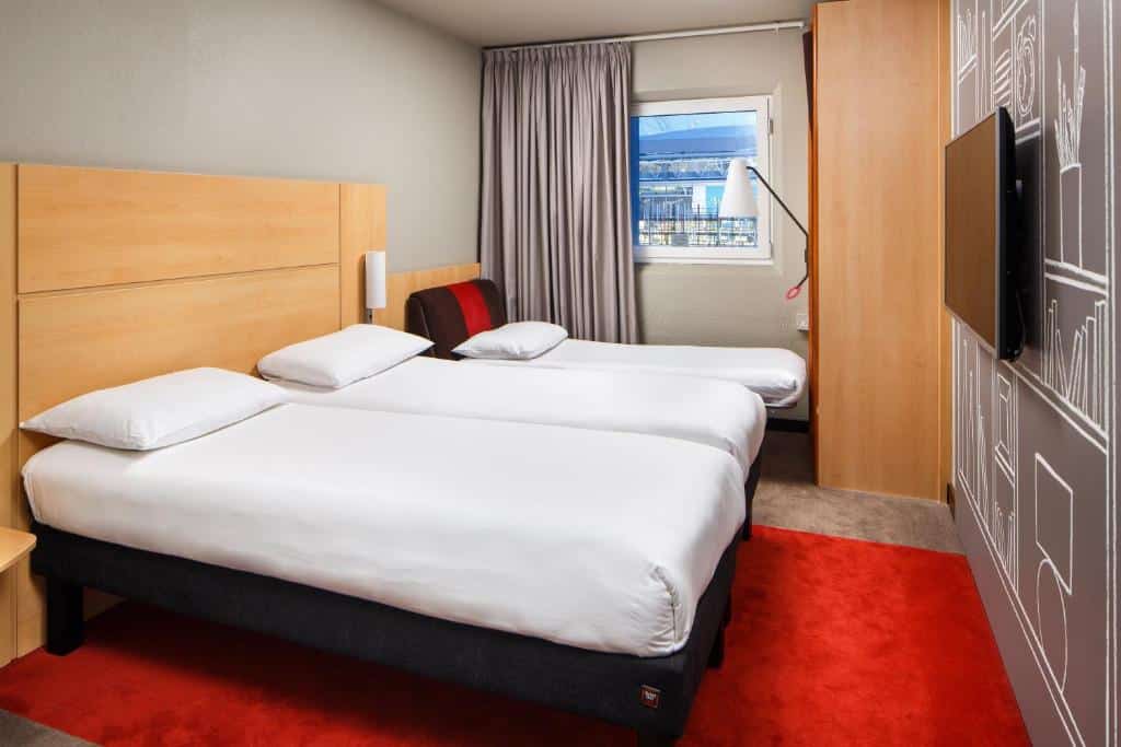 Quarto do ibis London Wembley com uma janela com cortinas, uma cama de solteiro e uma de casal, há uma televisão e um tapete vermelho, para representar hotéis Ibis em Londres