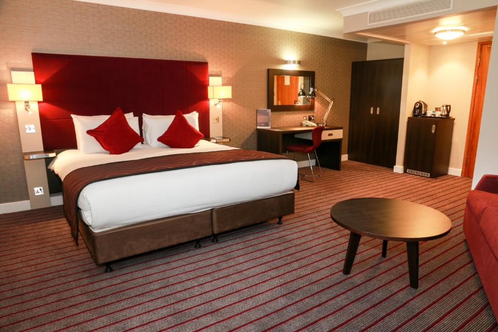Quarto do Mercure London Heathrow Airport com uma cama de casal, uma mesa de escritório com uma cadeira, um armário pequeno, um frigobar, um carpete cinza e vermelho, além de um sofá, para representar hotéis Mercure em Londres