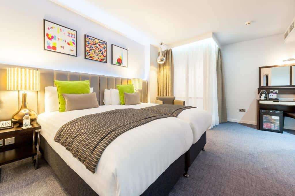 Quarto do Mercure London Paddington Hotel com duas camas de solteiro, carpete cinza, janela com cortinas, um frigobar e dois abajures nas laterais das camas, há também uma poltrona