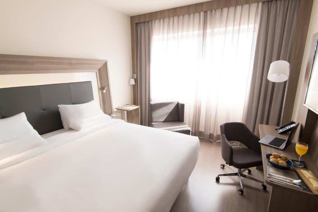 Quarto do Novotel São Paulo Berrini com uma cama de casal, uma janela com cortinas, uma poltrona, uma mesa de escritório com uma poltrona giratória, para representar hotéis para o The Town em SP