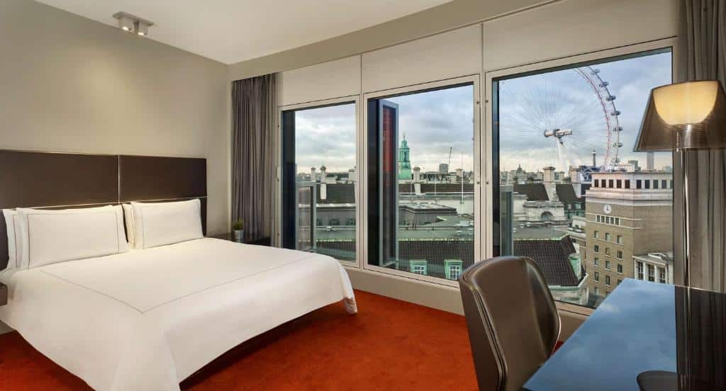 Quarto do  Park Plaza Westminster Bridge London com três janelas grandes com vista para a cidade e a London Eye, uma cama de casal, carpete vermelho, uma mesa de escritório com uma cadeira