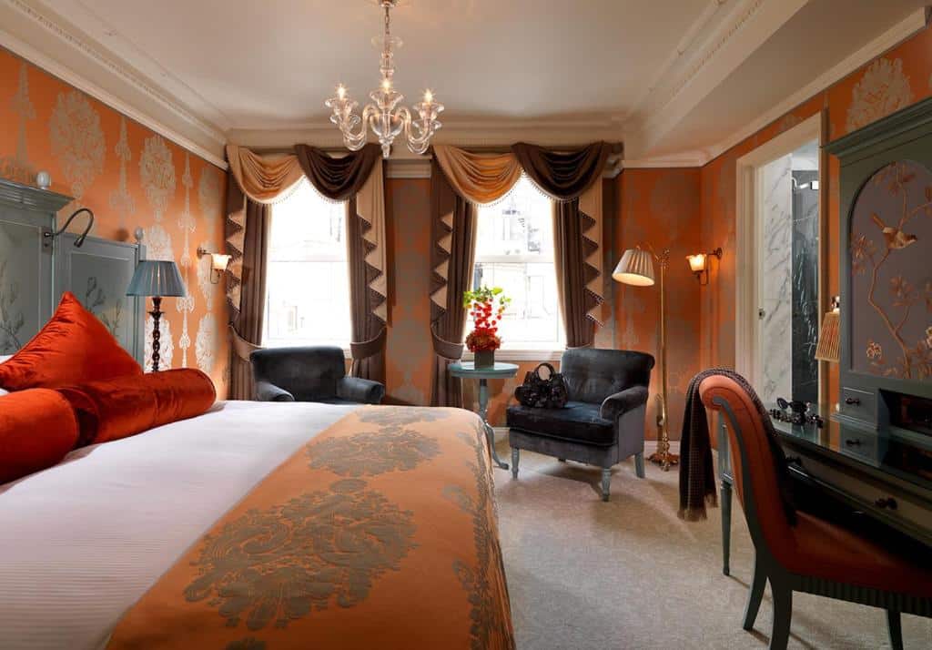 Quarto do The Goring com duas janelas com cortinas, uma lustre, papel de parede laranja com detalhes em prata, uma cama de casal, duas poltronas e uma mesa de escritório, além de um abajur de chão