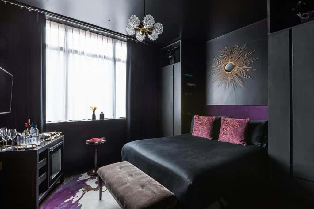 Quarto do The Mandrake inteiro preto com detalhes em vinho, há uma janela com cortinas, um lustre, uma televisão e um móvel com itens de bebidas, o chão é de carpete em tons de roxo e branco, para representar hotéis no centro de Londres