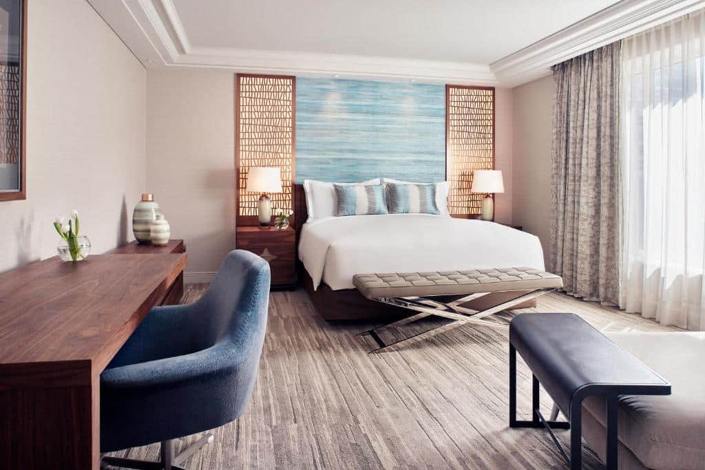 Quarto do The Ritz-Carlton, com cama de casal no centro do quarto, duas cômodas ao lado da cama com luminária e do lado esquerdo mesa de trabalho com cadeira.