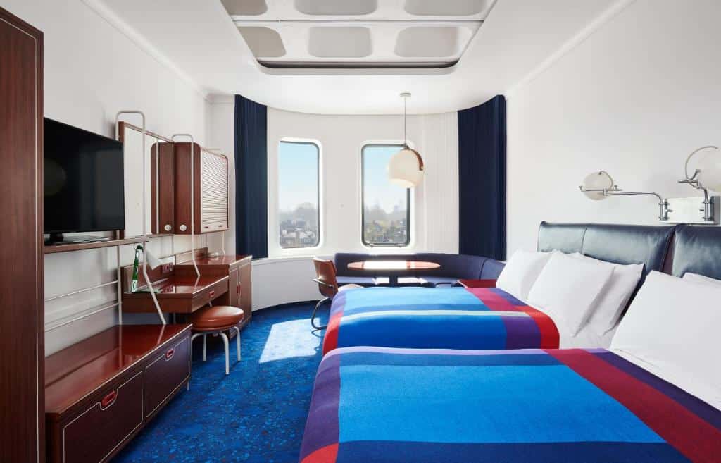Quarto do The Standard London, com duas janelas, uma cama de casal e uma de solteiro, há uma mesa de escritório e uma televisão, além de um sofá de canto. O ambiente é decorado em tons de azul, roxo, vermelho e madeira