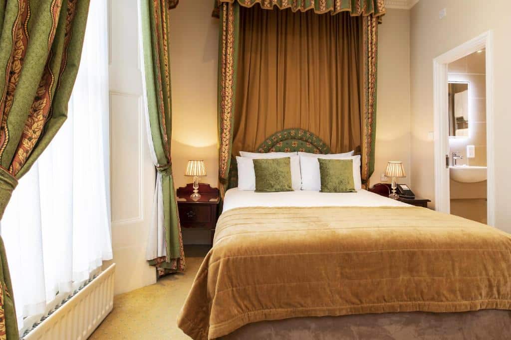 Quarto do The Windermere Hotel, London com uma cama de casal, uma cabeceira que vai até o teto com tecidos em tom de marrom e verde, uma janela com cortinas e duas mesinhas de cabeceira com abajures, para representar hotéis no centro em Londres