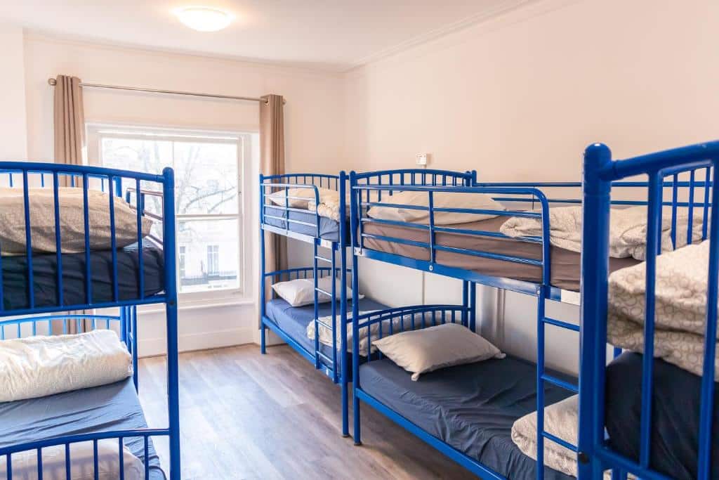 Quarto no Urbany Hostel London com várias beliches azuis com travesseiros, além de uma ampla janela com cortina e o chão é de madeira