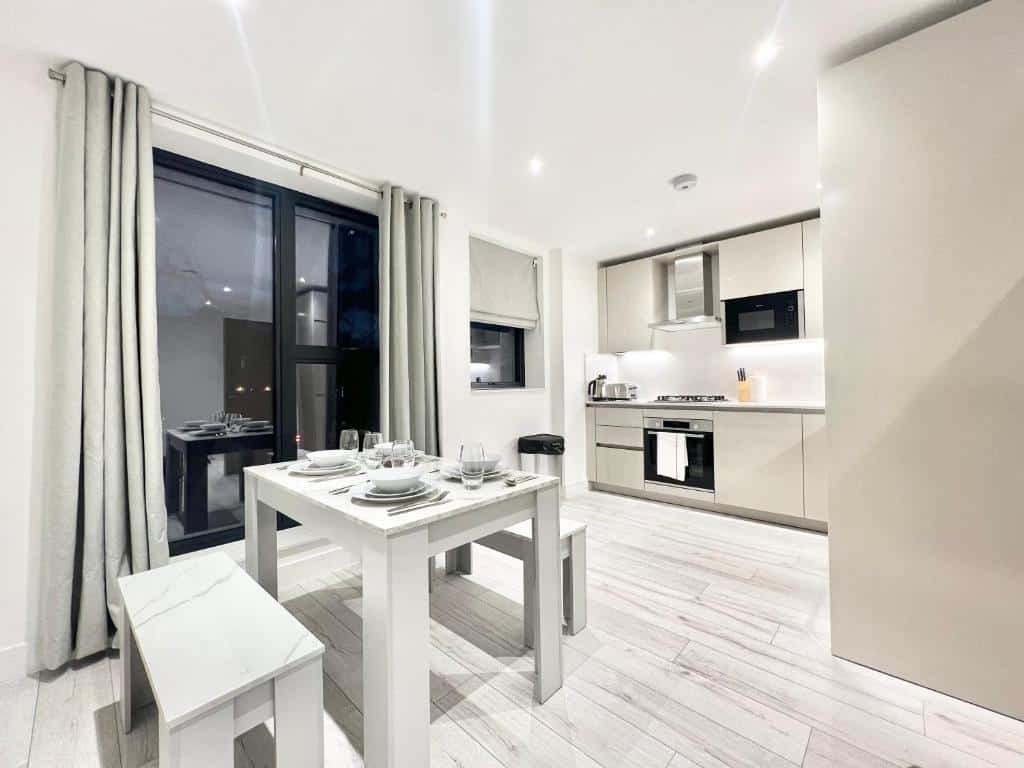 Cozinha do Queens Luxury Apartments com uma mesa com dois bancos, uma sacada com cortinas, chão de madeira clara, para representar aluguel de temporada em Londres