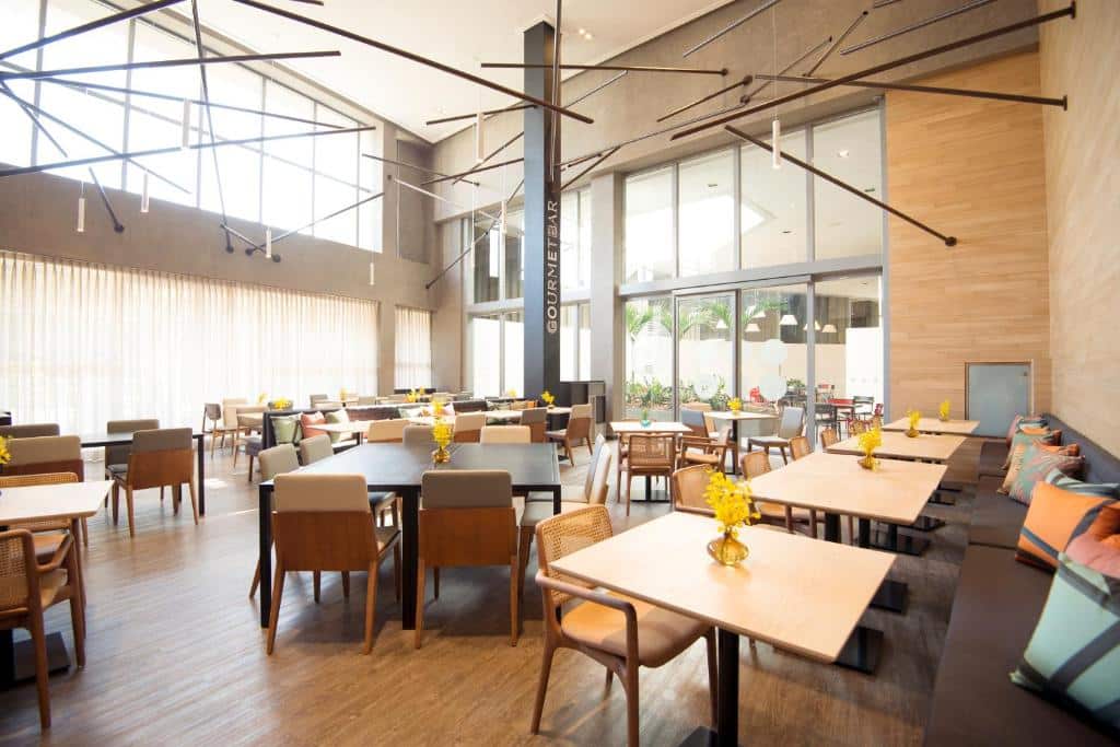Salão de refeições do Novotel São Paulo Berrini com teto alto, mesas quadradas, cadeiras de madeiras e sofás com almofadas coloridas