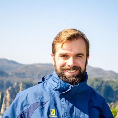 Foto do perfil de Rodrigo Belasquem, de barba e cabelos castanhos, usando blusa de frio até o pescoço azul, sorrindo, com uma paisagem composta por montanhas ao fundo
