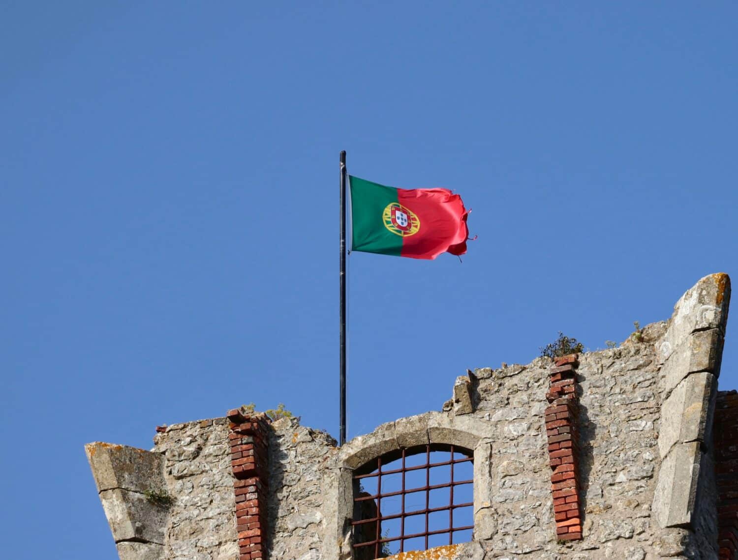 Uma bandeira de Portugal, vermelha e verde, presa ema um construção de pedras, para representar quanto custa seguro viagem Portugal