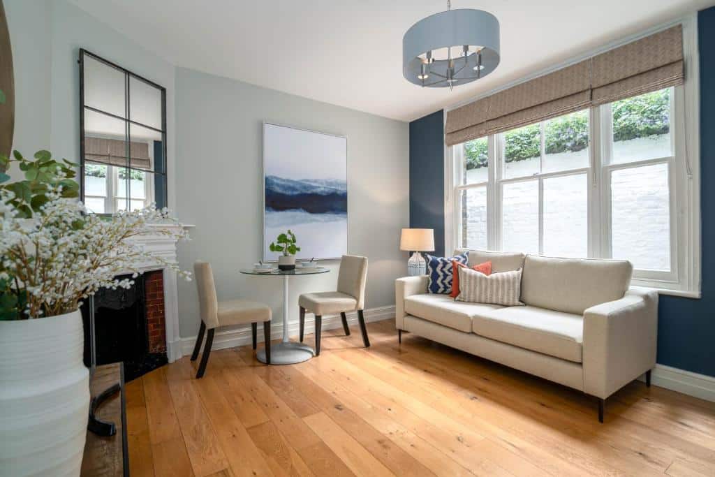 Sala de estar do 5 Doughty Street com chão de madeira, um sofá com almofadas, uma janela com persianas e uma pequena mesa com duas cadeiras, para representar airbnb em Londres