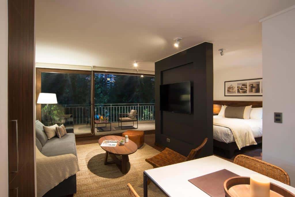 Vista da sala e quarto do The Place com sofá cinza, mesa de centro do lado esquerdo, do lado direito cama de casal. Representa hotéis em Providencia em Santiago.