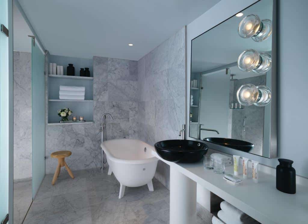 Banheiro amplo do Sea Containers London com uma banheira oval, pia espaçosa, espelho, prateleiras com itens de higiene e uma porta para o box, para representar hotéis bem localizados em Londres