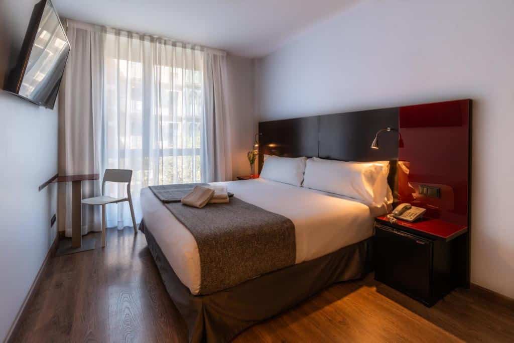 Quarto do SM Hotel Sant Antoni, uma das recomendações de hotéis perto da Sagrada Família em Barcelona. A cama de casal tem um painel de madeira atrás e mesinhas de cabeceira com luminária dos dois lados. Há uma televisão de frente para a cama. A janela está coberta por cortinas bege e tem uma mesa com cadeira ao lado.