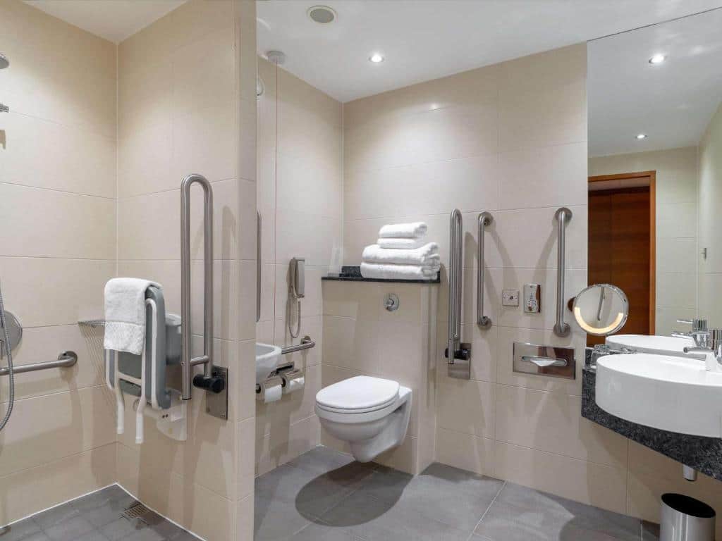 Banheiro adaptado do Sofitel London Heathrow com barras de apoio, box sem vidro e aberto, e toalhas espalhadas por todos os lados, para representar hotéis perto do Aeroporto de Londres Heathrow