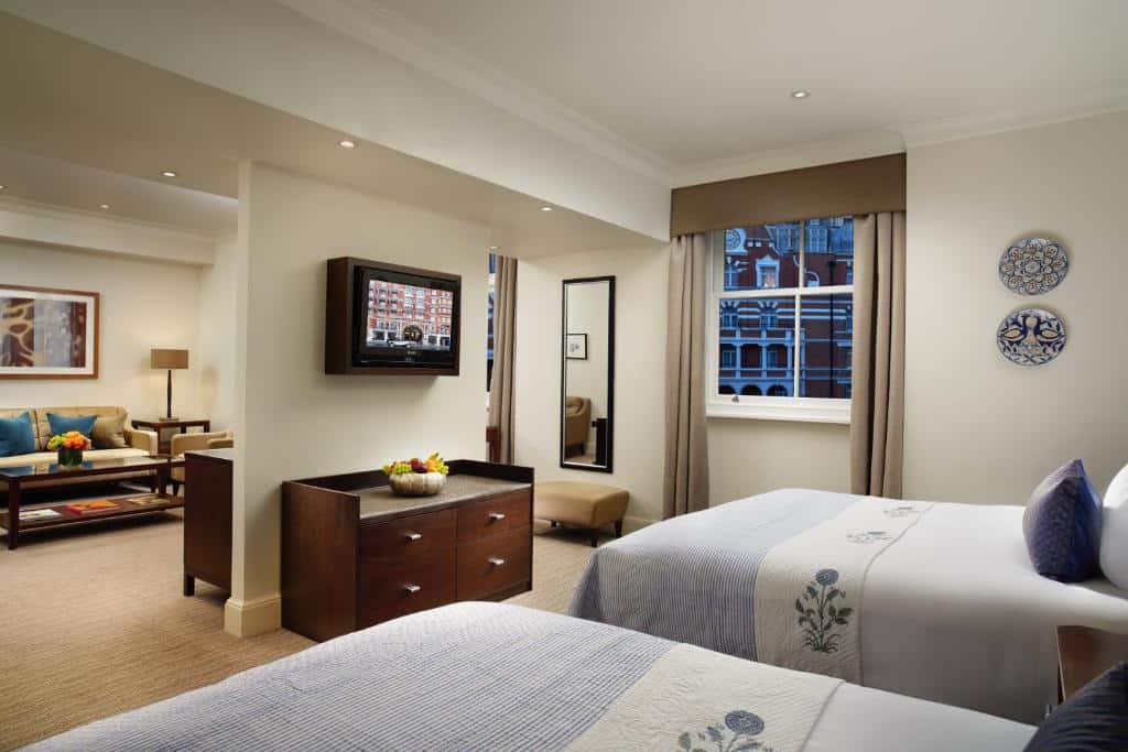 Quarto do St. James’ Court, A Taj Hotel, London com duas camas de casal, uma janela com cortinas, um espelho de corpo inteiro, há uma sala conjugada com sofás, rack para televisão e um abajur