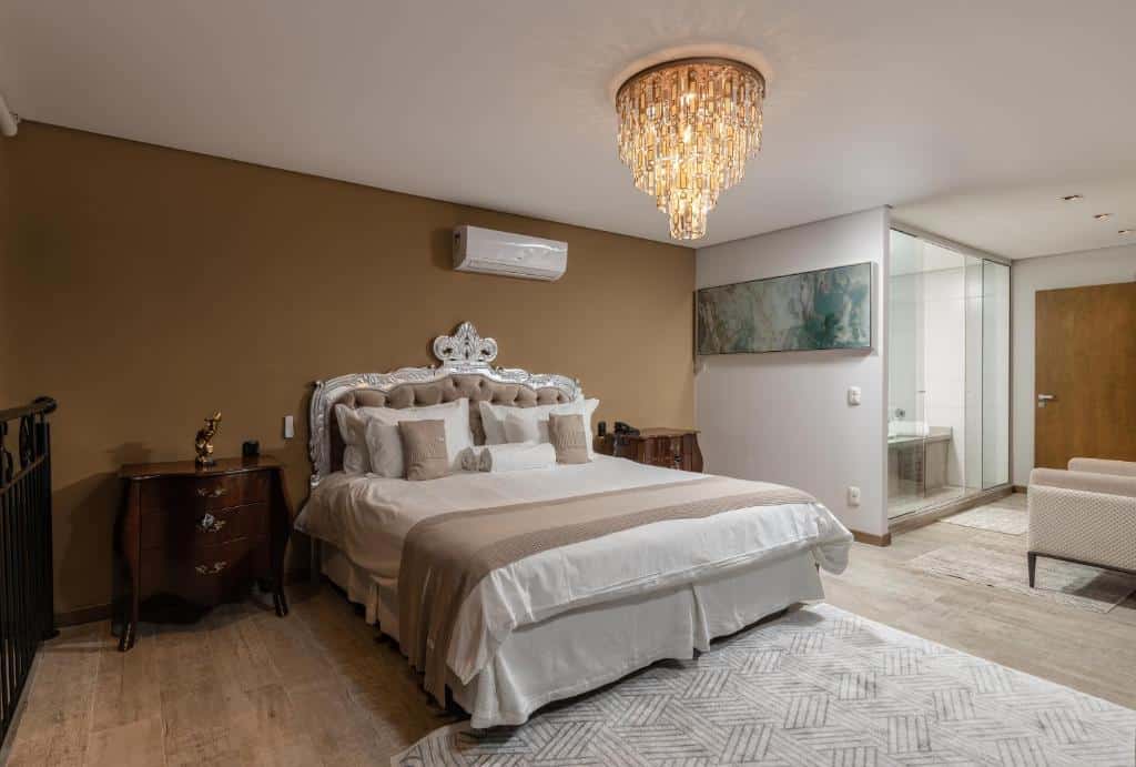 quarto do Hotel Fioreze Centro com uma cama de casal elegante no centro, uma porta de vidro mostrando uma banheira no canto direito e um lustre de vidro aceso no teto