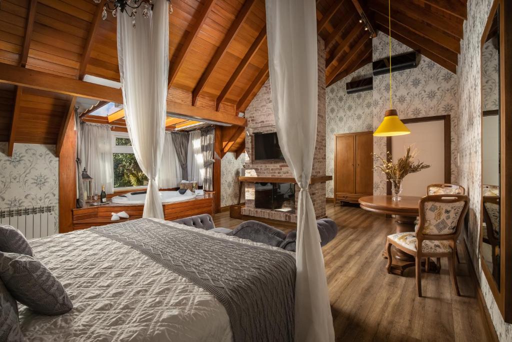 suíte do Hotel Valle Dincanto em Gramado com uma banheira de hidromassagem, uma lareiro no meio do quarto, uma mesa de madeira com duas cadeiras estofadas no lado direito e uma cama de casal no canto inferior esquerdo.
