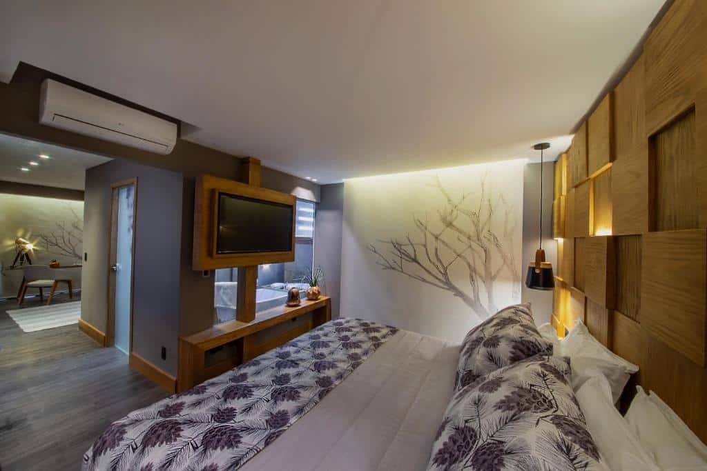 suíte do Wood Hotel em Gramado com uma cama de casal no canto inferior direito, uma banheira de hidromassagem em frente a cama e uma área de estar mais afastada, no lado esquerdo da imagem