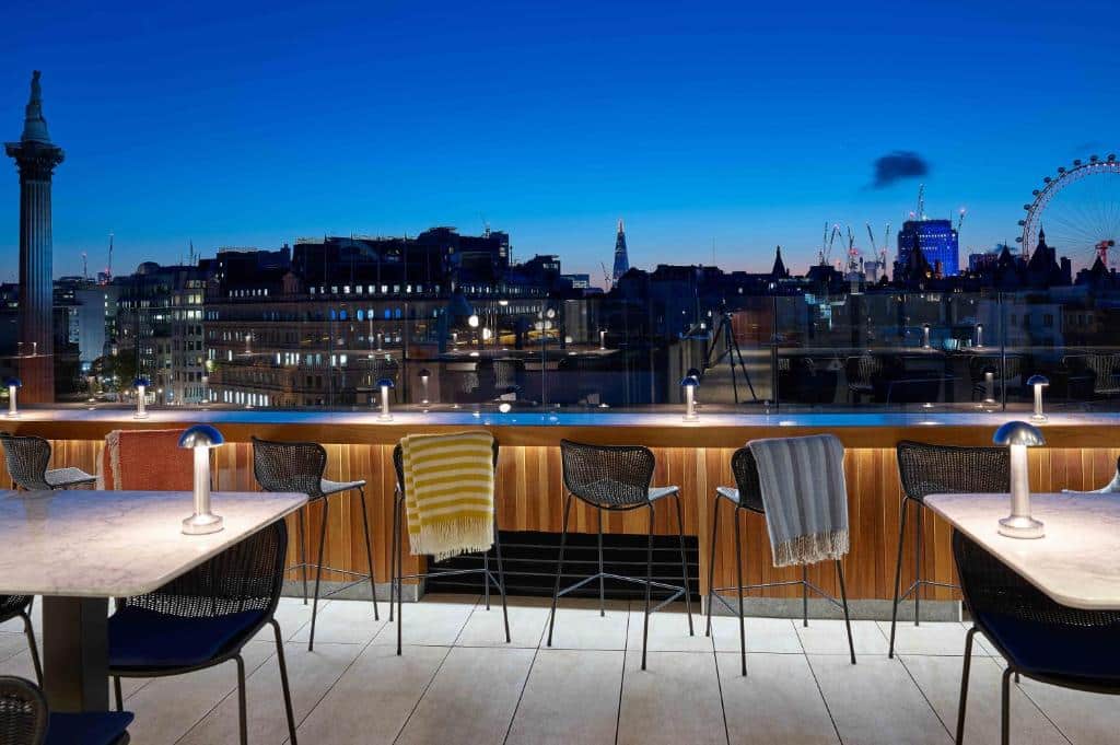 Varanda do The Trafalgar St. James, London Curio collection by Hilton com umas cadeiras com cobertas, uma mesinha com cadeiras, e há uma proteção de vidro com vista para a cidade