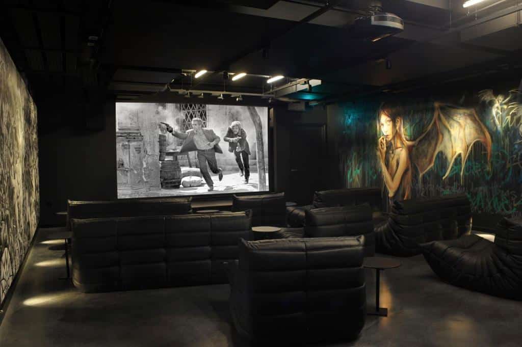 Sala de cinema com poltronas e sofás no The Mandrake com paredes cheias de desenhos e uma tela de cinema, para representar hotéis no centro de Londres