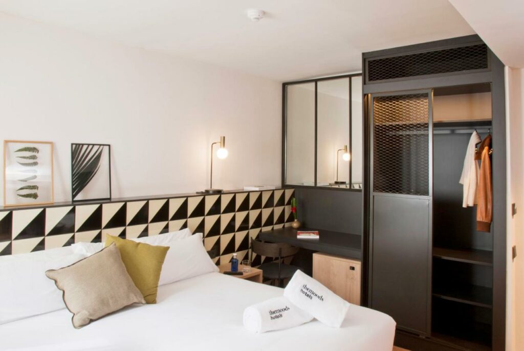 Quarto do The Moods Oasis, uma das recomendações de hotéis baratos em Barcelona.  A cama de casal está encostada em uma cabeceira branca e preta e tem mesinhas de cabeceira dos dois lados com luminária. Há duas toalhas dobradas em cima da cama. Ao lado há uma mesa com cadeira e ármario de roupas com prateleiras.
