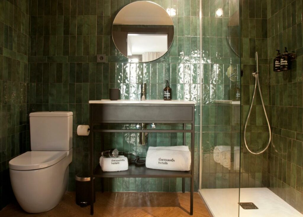 Banheiro do The Moods Oasis. Uma privada branca está ao lado da pia, que tem um suporte com toalhas e secador de cabelo. Há um espelho na parede acima da pia. Ao lado, um chuveiro está protegido por box de vidro. O banheiro é de azulejo verde.