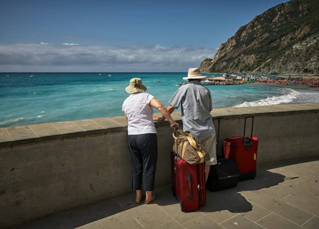 Um casal de idosos com malas de viagem observando o mar de uma praia, para representar quanto custa seguro viagem Portugal