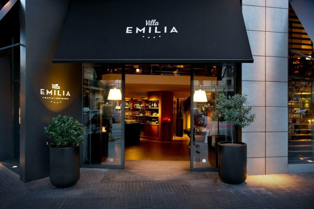 Entrada do Hotel Villa Emilia, um dos melhores hotéis em Barcelona. O nome do hotel está escrito na parede ao lado e no toldo preto acima. Há uma planta de cada lado da entrada em vasos pretos. Lá dentro a iluminação é amarelada.