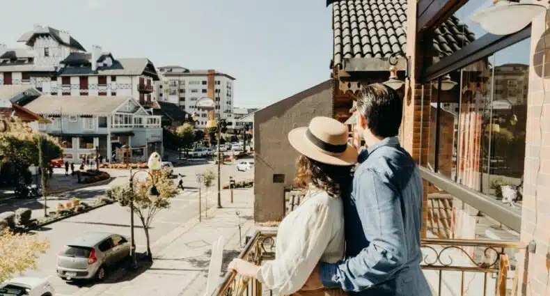 um casal abraçado, no lado direito da imagem, olhando para rua de Gramado de dentro de uma sacada aberta. Há carros na rua abaixo, no canto esquerdo inferior