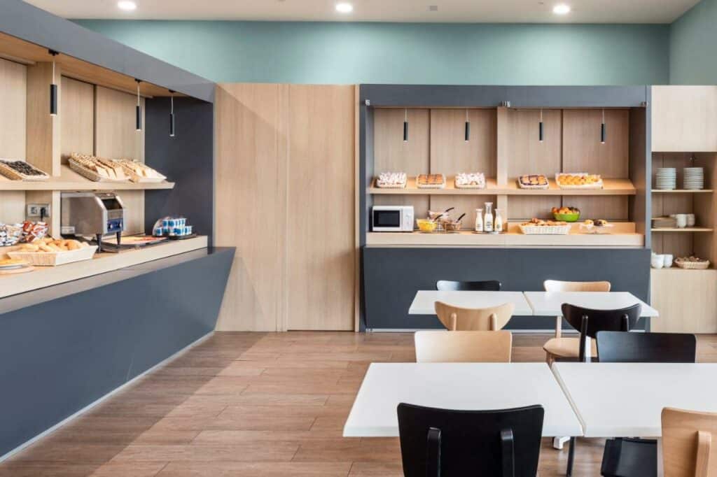 Local de café da manhã do B&B HOTEL Barcelona Viladecans. Balcões com frutas, pães e frios estão ao redor. Há um microondas e uma torradeira, e do lado direito um suporte está com pratos e outros utensílios. Há mesas e cadeiras dispostas no local.