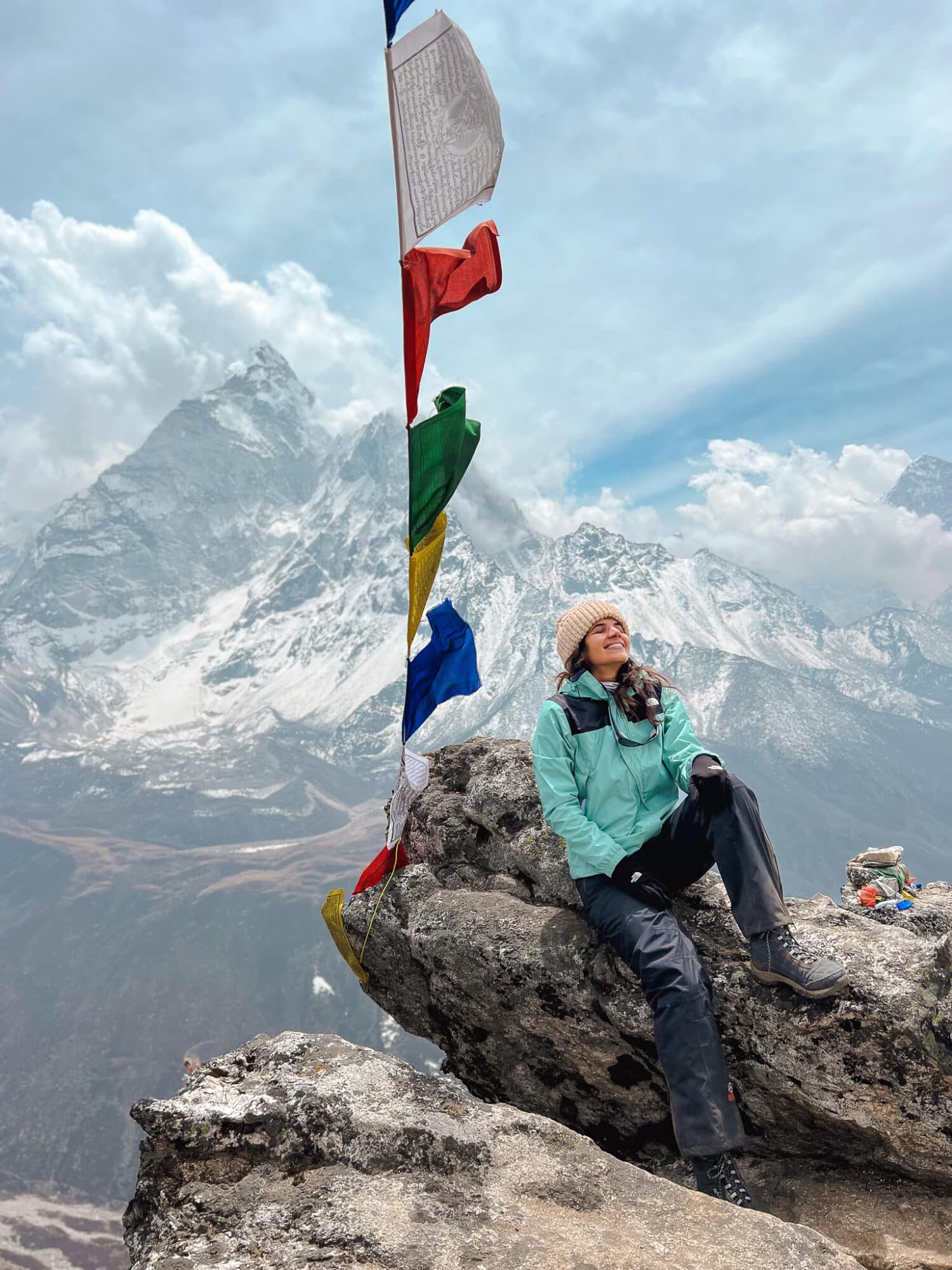 Mulher sentada em uma pedra, com os olhos fechados e a cabeça levantada, com várias bandeiras coloridas ao lado e, ao fundo, várias montanhas de gelo