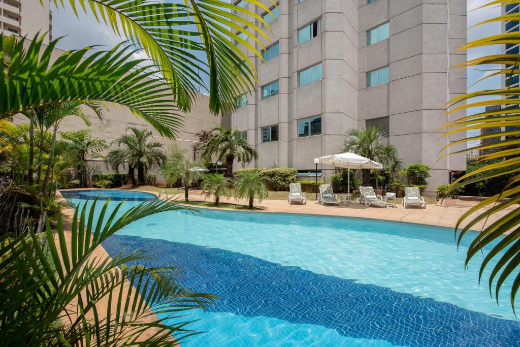 Longa piscina externa no hotel Intercity São Paulo Ibirapuera. Há várias palmeiras ao redor da piscina e também há espreguiçadeiras com guarda-sol.