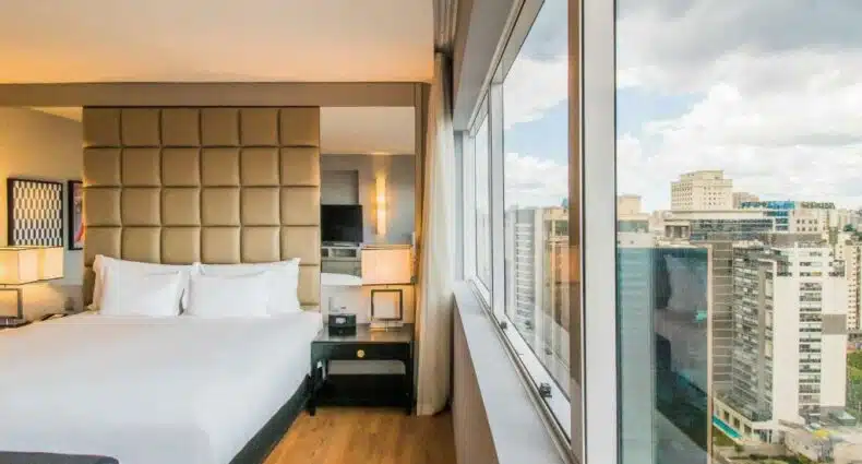 Vista de uma das suíte do hotel Pullman São Paulo Vila Olímpia. É possível ver uma cama box de casal com lençóis e dois travesseiros brancos no lado esquerdo da foto. Há uma mesa de cabeceira preta com gaveta do seu lado, comportando um abajur e outros itens. Atrás da cama, há uma cabeceira em estilo quadriculado na cor bege, com dois espelhos do lado. Do lado direito da foto, está a janela com visão para a cidade de São Paulo. Foto para representar o post sobre hotéis em Vila Olímpia SP.