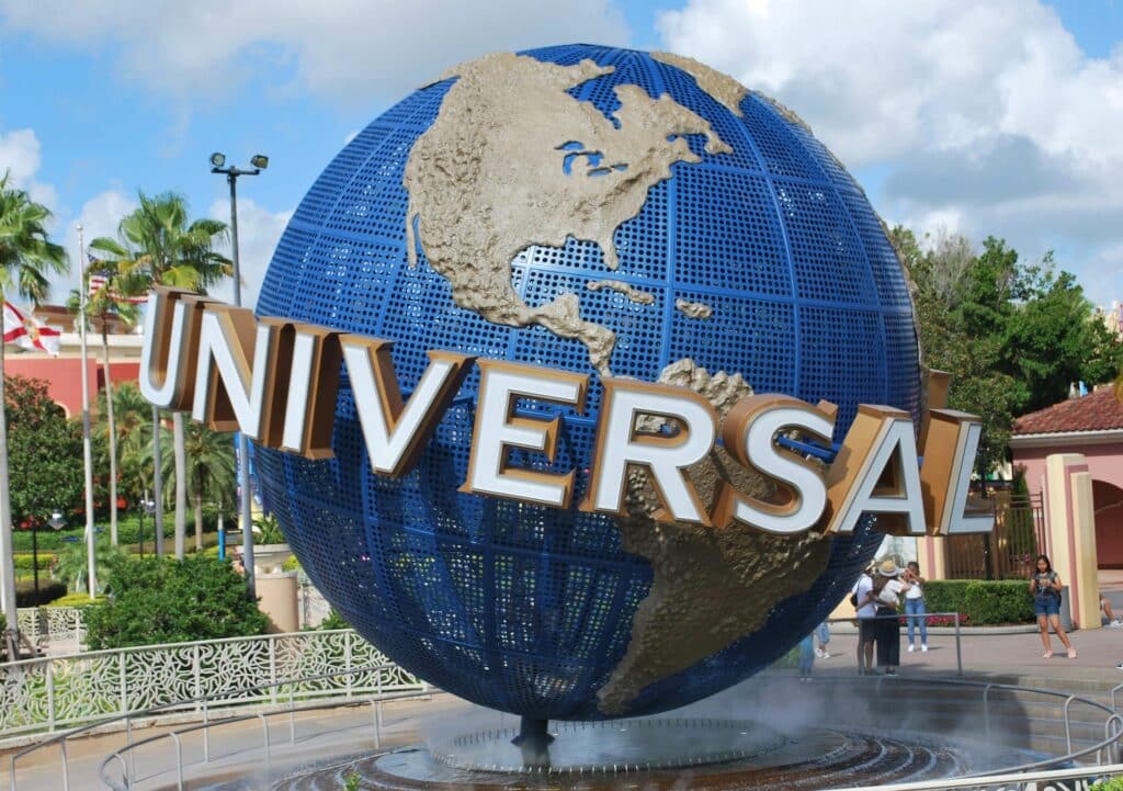 vista do Universal Studios Plaza, em Orlando, um globo terrestre com os escritos UNIVERSAL grande em frente, o céu está azul com nuvens e há algumas pessoas caminhando ao redor