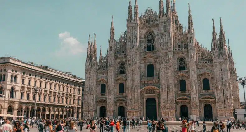 Praça em frente a Catedral de Milão com diversas pessoas andando, a catedral é construída em estilo gótico e com diversas pequenas torres
