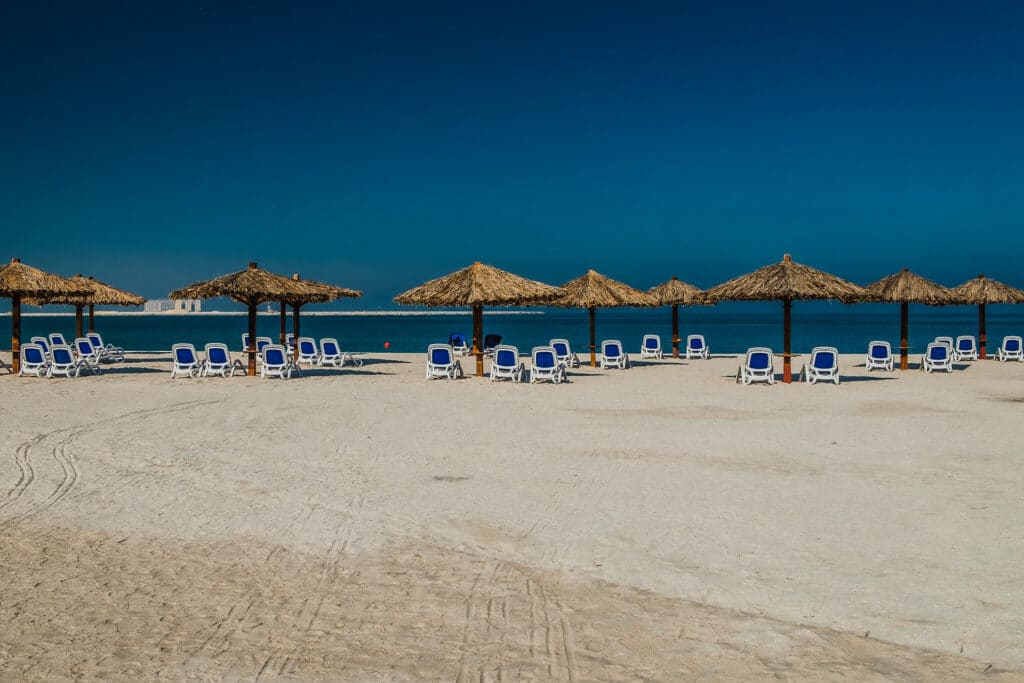 Praia de Al Jazeerah com uma extensa faixa de areia com guarda-sóis de palha e cadeiras azuis e brancas embaixo deles, para representar o que fazer em Ras Al Khaimah