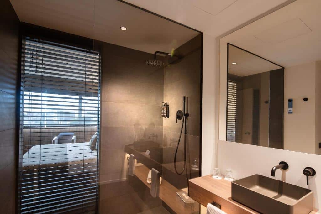 Banheiro do BAH Barcelona Airport Hotel. A porta do box de vidro reflete um pedaço do quarto, mas é possível ver chuveiro e amenidades de banho ali. Do lado de fora está a pia com dois copos de vidro e um espelho na parede acima.