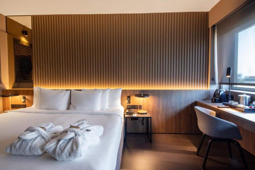 Quarto do BAH Barcelona Airport Hotel uma das recomendações de hotéis perto do aeroporto de Barcelona. A cama de casal tem mesinhas de cabeceira dos dois lados com luminárias, e dois roupões estão dobrados aos pés da cama. Do lado direito está uma mesa de trabalho com cadeira, abajur e amenidades de café e chá. A mesa encara uma janela.