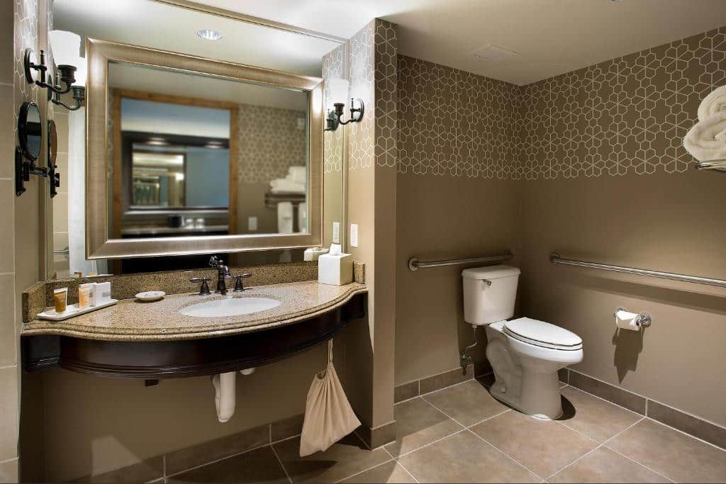 Banheiro espaçoso e adaptado do Hotel Contessa – Suites on the Riverwalk com barras de apoio nos arredores do vaso sanitário e uma pia mais baixa