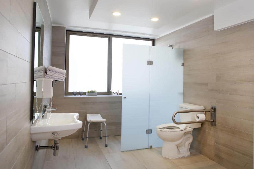 Banheiro com acessibilidade do MR Express (ex Hotel Neruda Express) com vaso sanitário do lado direito com barra de apoio, em frente pia baixa e do lado esquerdo do ambiente chuveiro com cadeira.