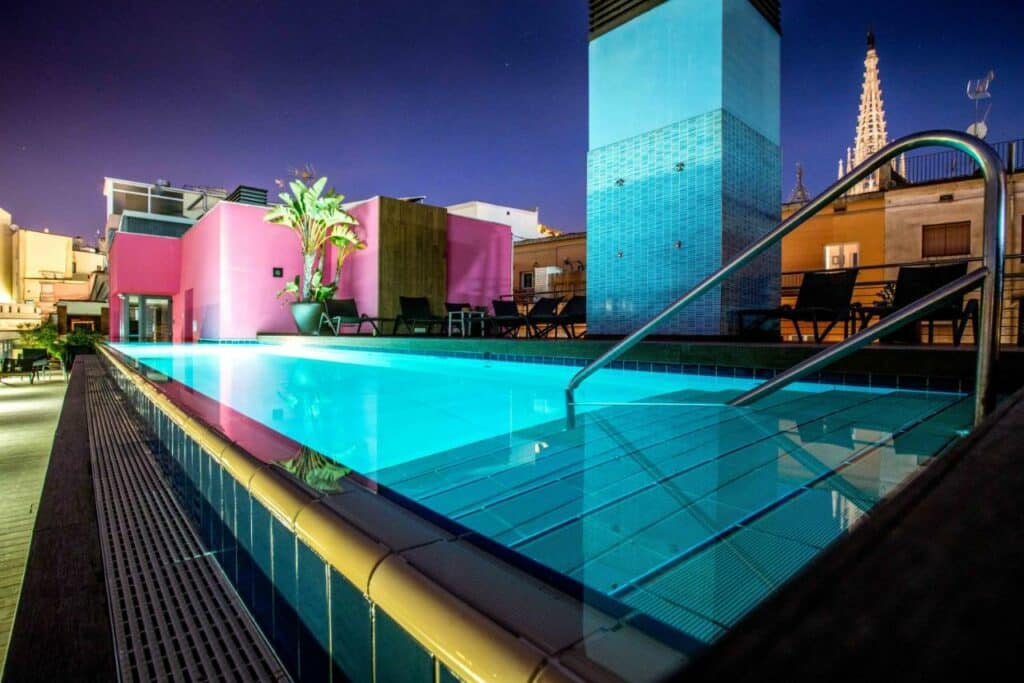 Piscina do Hotel Barcelona Catedral, uma das das recomendações de hotéis no Bairro Gótico em Barcelona. Com iluminação aquática, a piscina tem espreguiçadeiras ao redor. As paredes cor de rosa do hotl podem ser vistas ao fundo com um vaso de plantas ao lado.