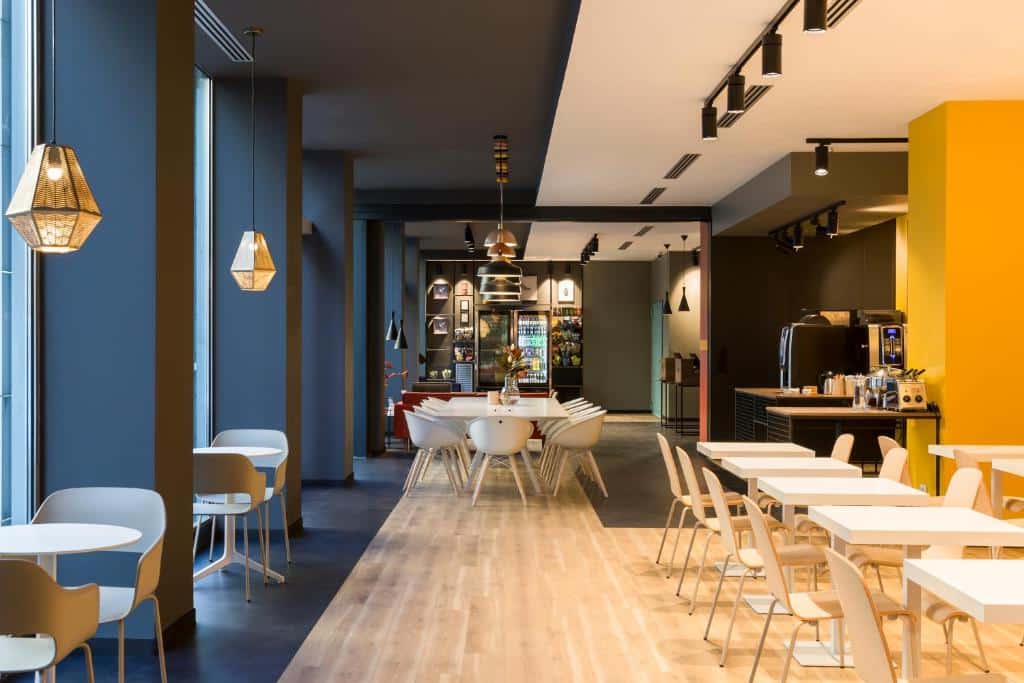 Salão para refeições do B&B Hotel Milano Central Station com mesas quadradas, cadeiras, luminárias presas no teto e um balcão com máquinas de café