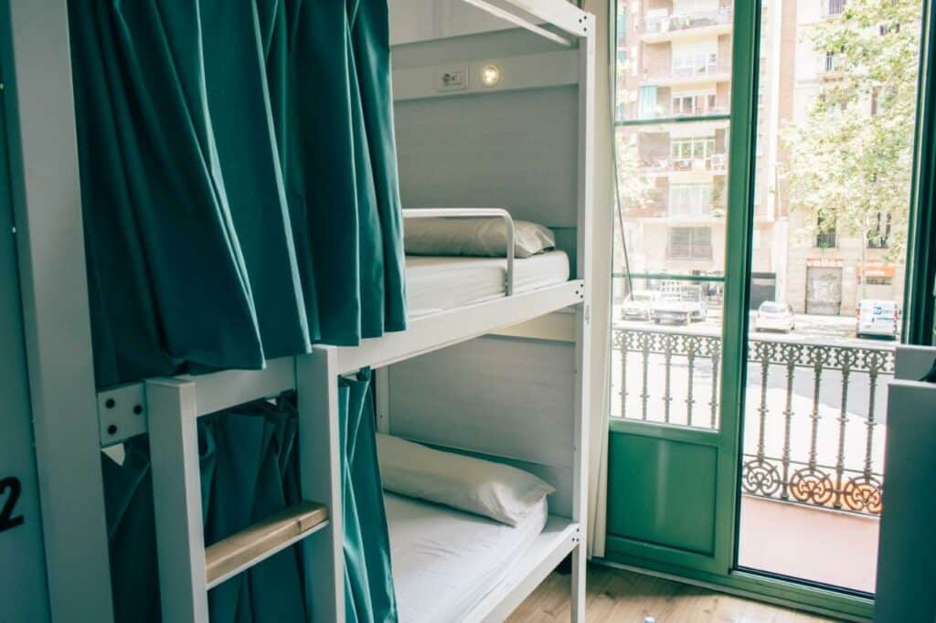 Quarto do Black Swan Hostel, uma das recomendações de hostels em Barcelona. Uma beliche numerada com cortina de privacidade está ao lado de uma porta de vidro com vista para a rua.