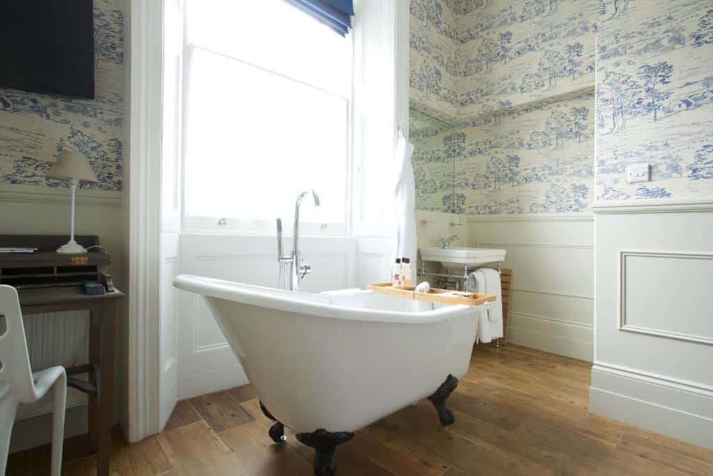 Uma banheira em um dos quartos do Bower House do lado de uma janela com cortinas, o chão é de madeira e há uma pia próxima com toalhas penduradas, para representar hotéis baratos em Londres