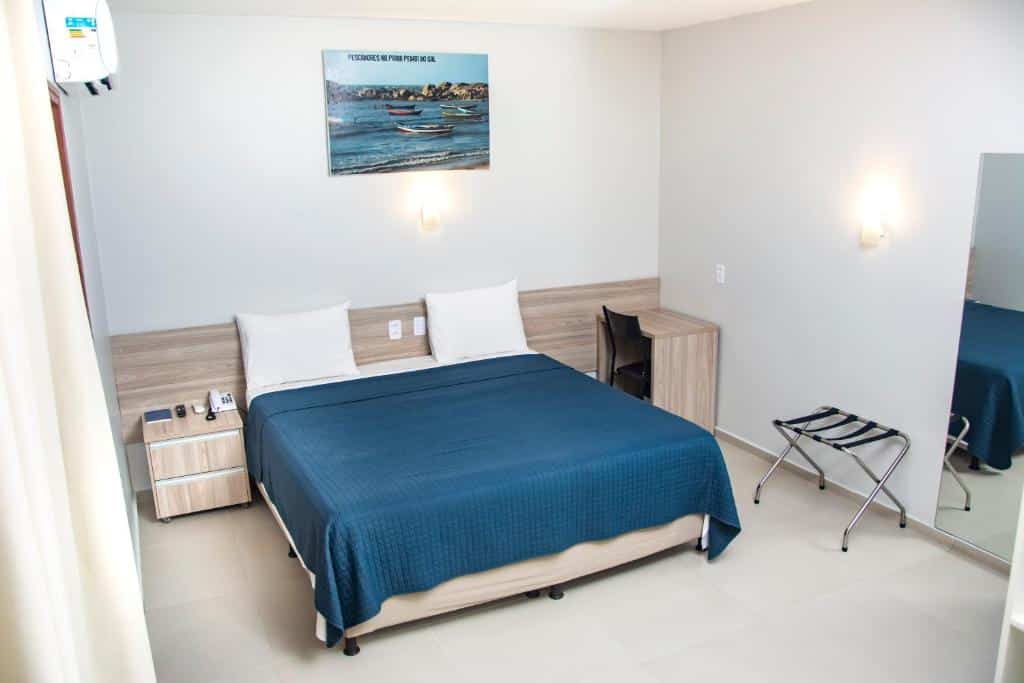 Quarto de hotel com decoração minimalista, cama de casal, pequenos móveis com madeira clara e paredes brancas. Imagem para ilustrar o post hotéis em Parnaíba.