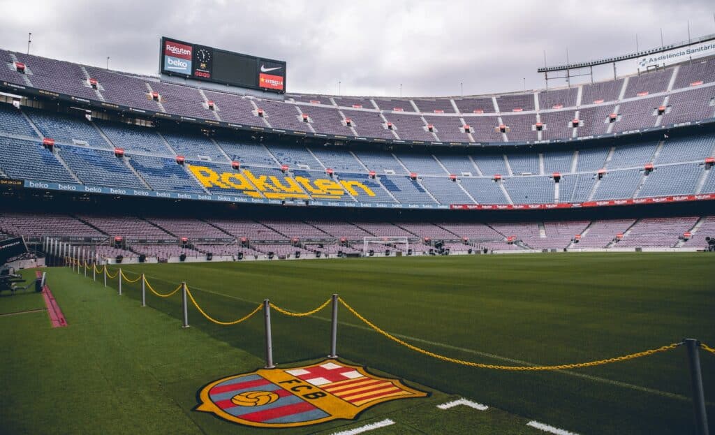 Estádio Camp Nou durante o dia. O escudo do Barcelona está estampado no chão perto de uma corrente que separa o campo. Ao fundo, as cadeiras do estádio estão vazias. - Foto: Patrick T'Kindt via Unsplash